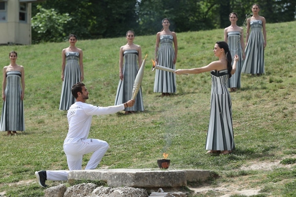 扮演最高女祭司的演員瑪麗·米娜（前右）點(diǎn)燃第一棒火炬手——希臘運(yùn)動(dòng)員、東京奧運(yùn)會(huì)男子賽艇項(xiàng)目金牌得主斯特凡諾斯·恩圖斯科斯手中的火炬。新華社記者 趙丁喆 攝